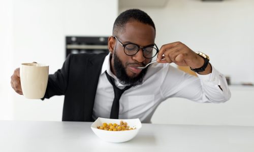 Modern Busy Lifestyle. Portrait of African American male in eyeglasses having breakfast in kitchen at home, wearing suit and drinking coffee while eating cereal, rushing to office, hurry to work